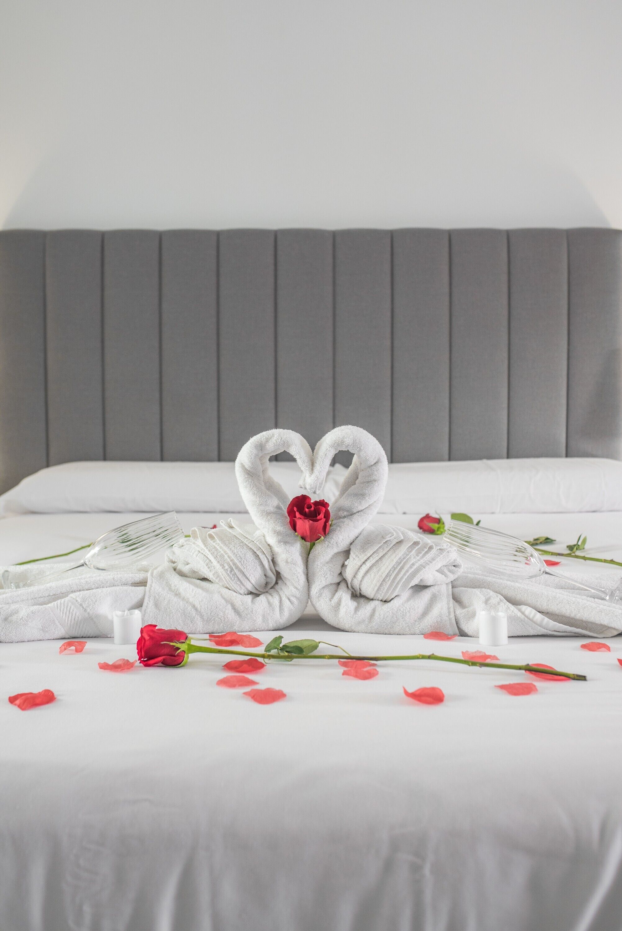 Un lit avec une tête de lit grise, du linge de lit blanc et une décoration en forme de cygnes, réalisée avec des serviettes, agrémentée d’une rose.