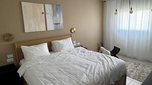 Un dormitorio con cama de madera, ropa de cama blanca, dos obras de arte en la pared, una mesita de noche y un buró, una ventana con cortinas traslúcidas, y una lámpara en el techo.
