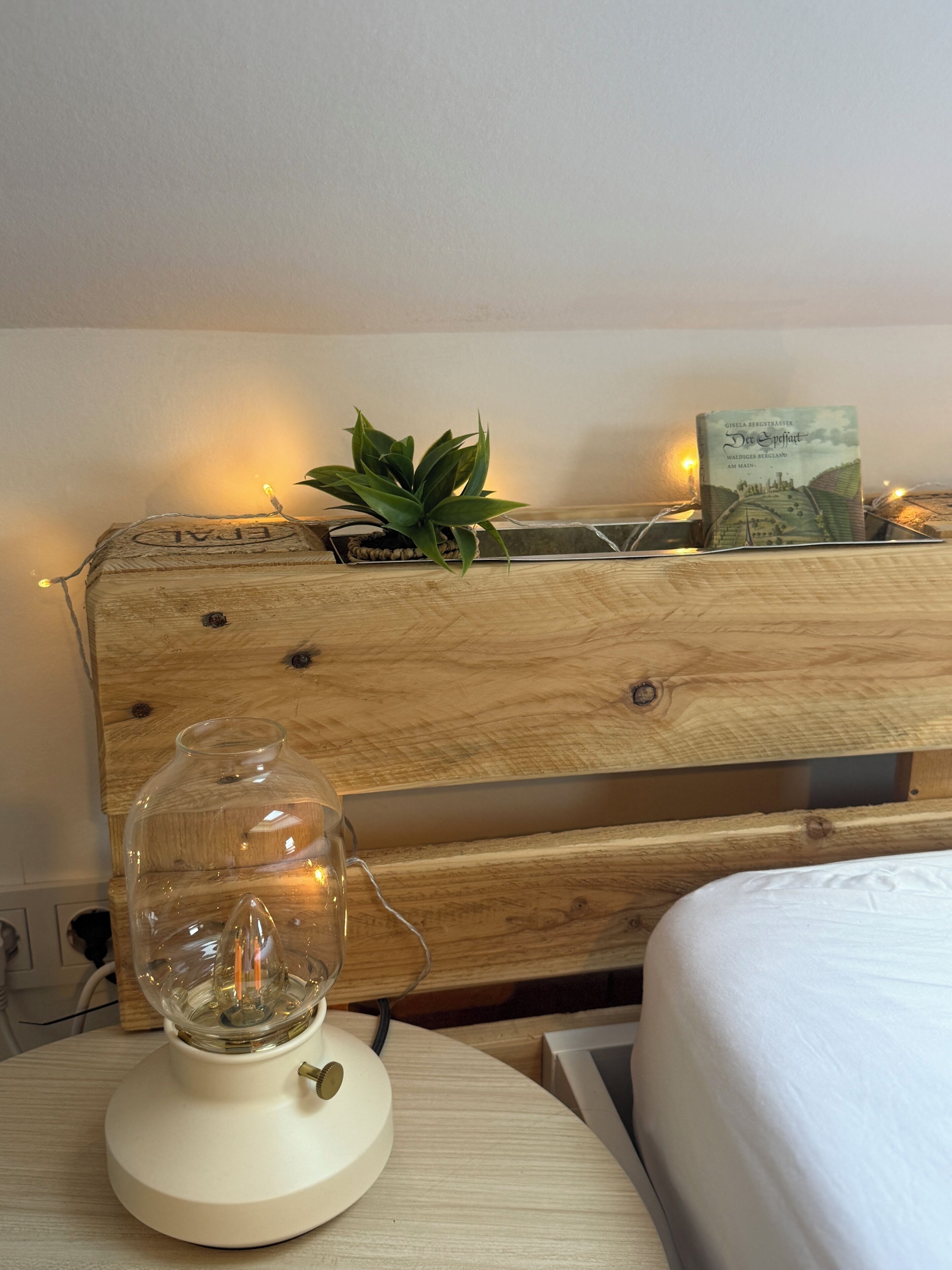 Cabecera de madera con una planta y un libro, una lámpara de cristal sobre una mesita de noche y una cama con ropa de cama blanca.