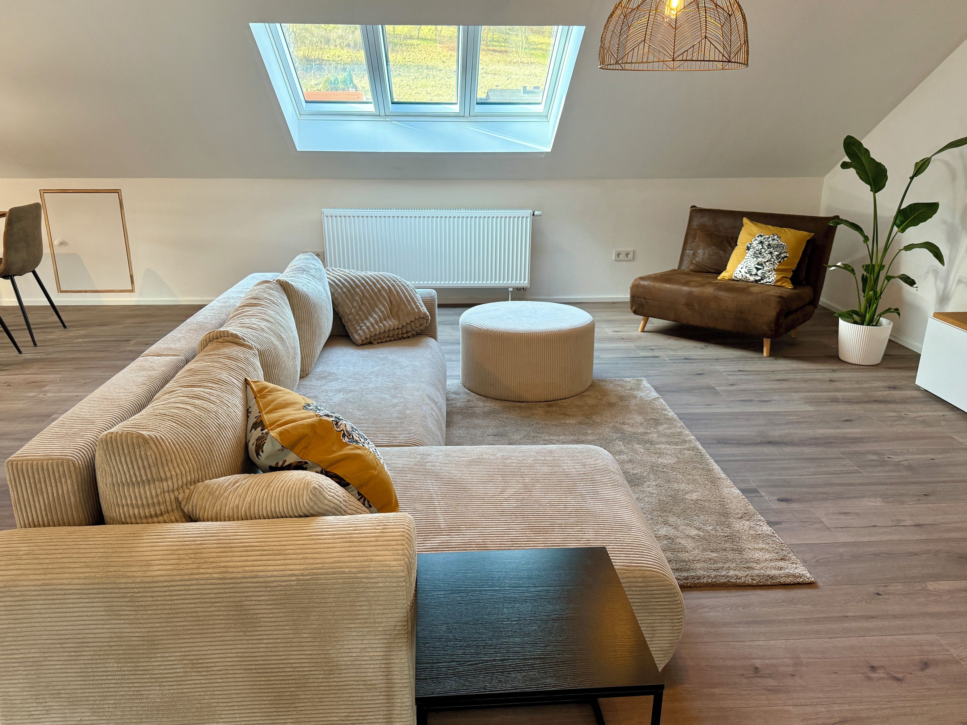 Een moderne woonkamer met een hoekbank, een poef en een salontafel. Er staat een bruine leren fauteuil met een kussen, een potplant en een dakraam erboven.