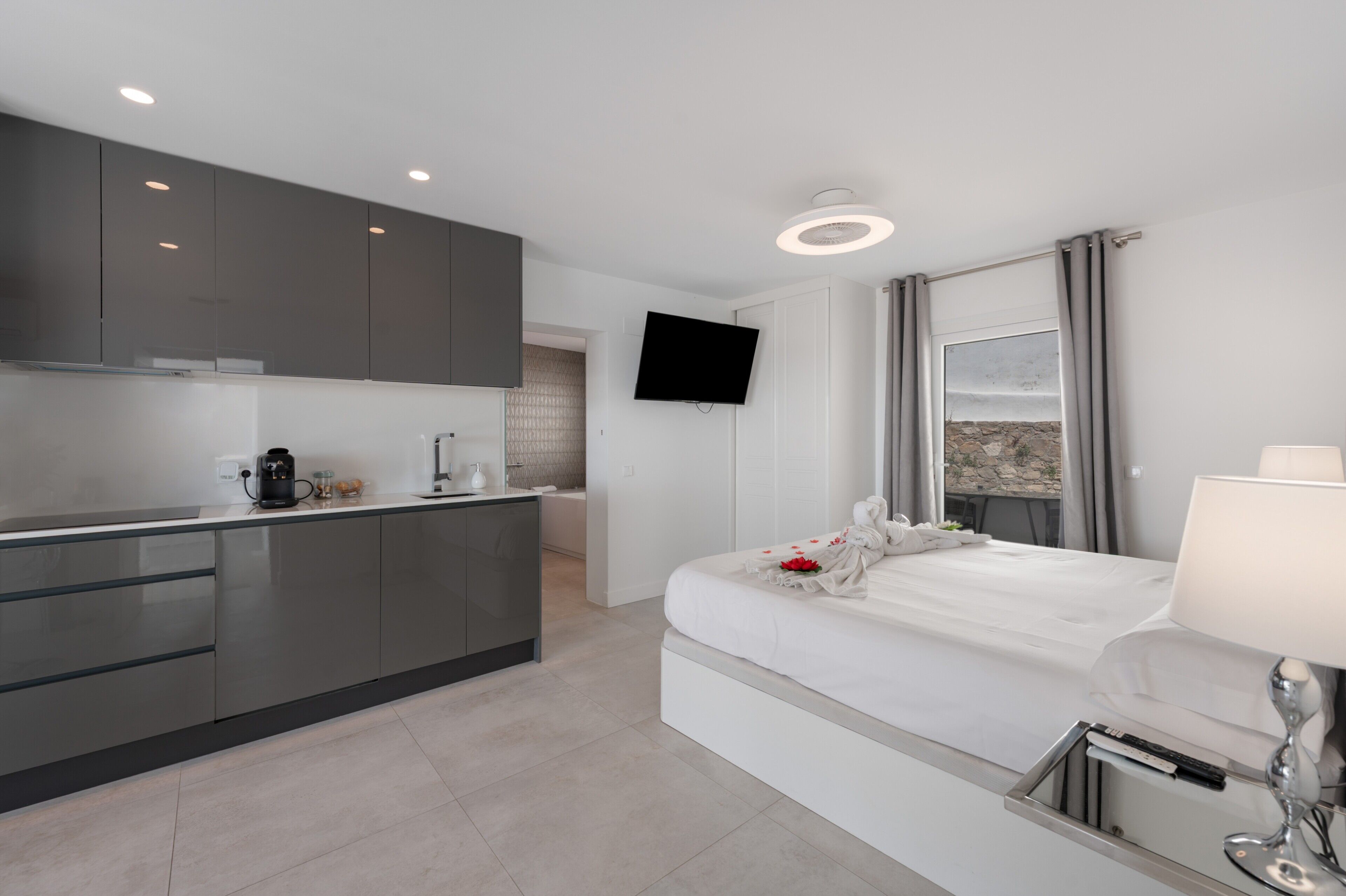 Una habitación de hotel moderna con kitchenette, televisión de pantalla plana, cama con cubrecama blanco y vistas al exterior.