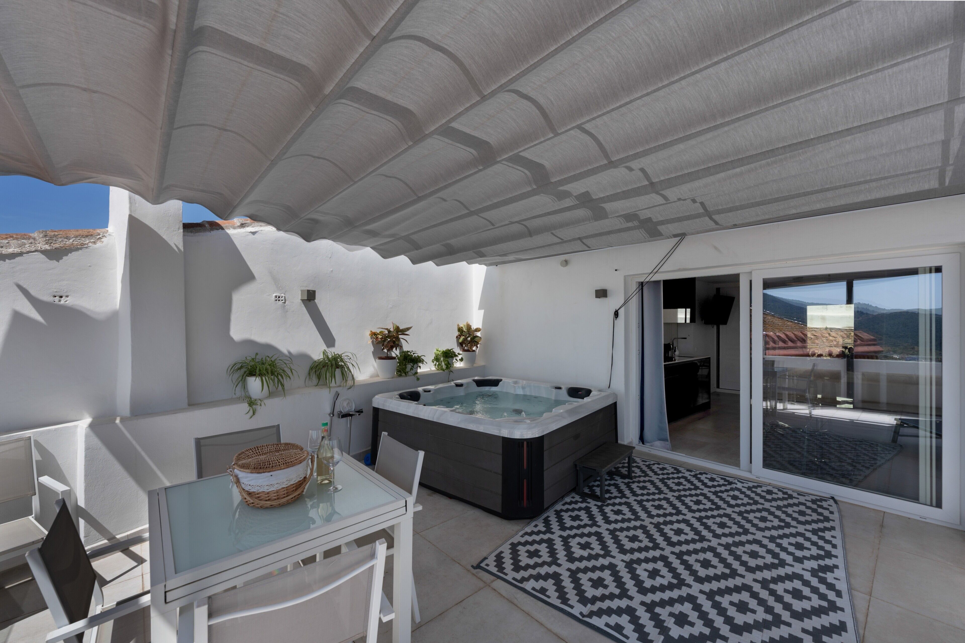 A modern outdoor living area with a glass table, chairs, a hot tub, and a patterned rug.