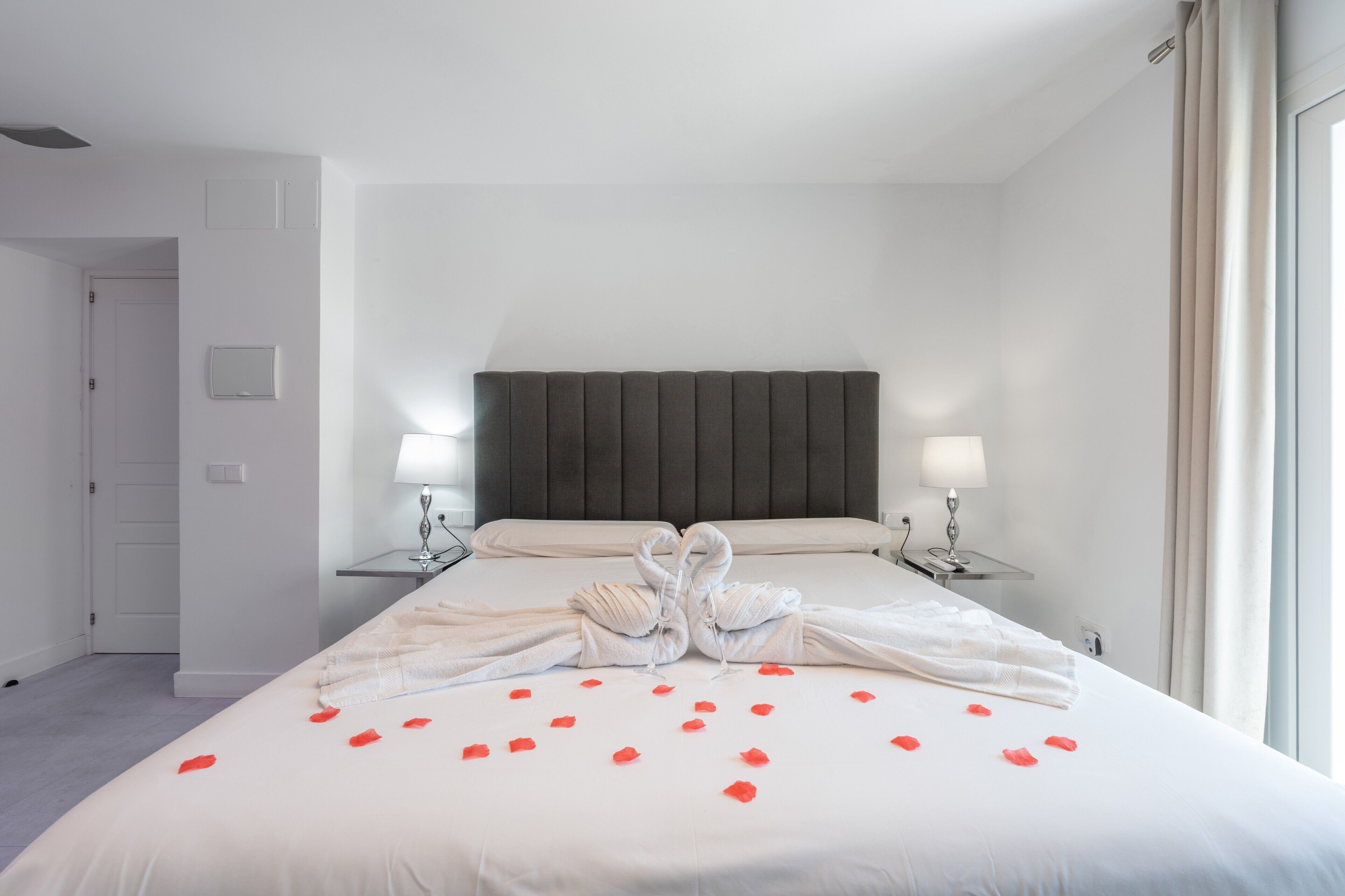 A hotel room with a large bed, two bedside lamps, a dark headboard, and a view of the outside through a window.