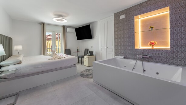 A modern bedroom with a large bed, a separate bathroom with a bathtub, a flat-screen TV mounted on the wall, and a small table with a chair. - Valeria Room: Double Whirlpool Bath, Mountain Views (Carratraca)