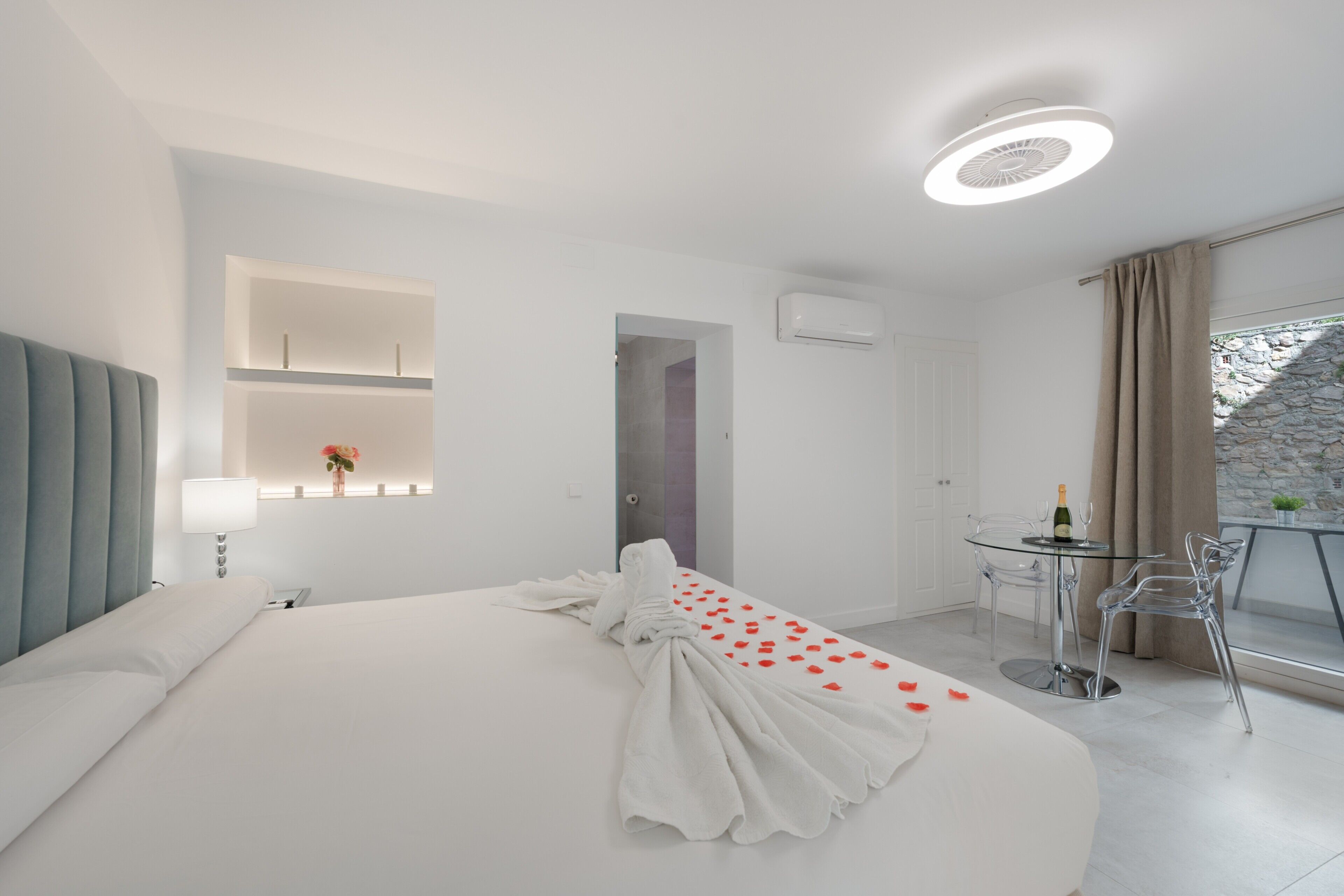 Una habitación de hotel moderna con una cama bien hecha, un pequeño comedor con sillas, una ventana con cortinas y una pared de piedra.