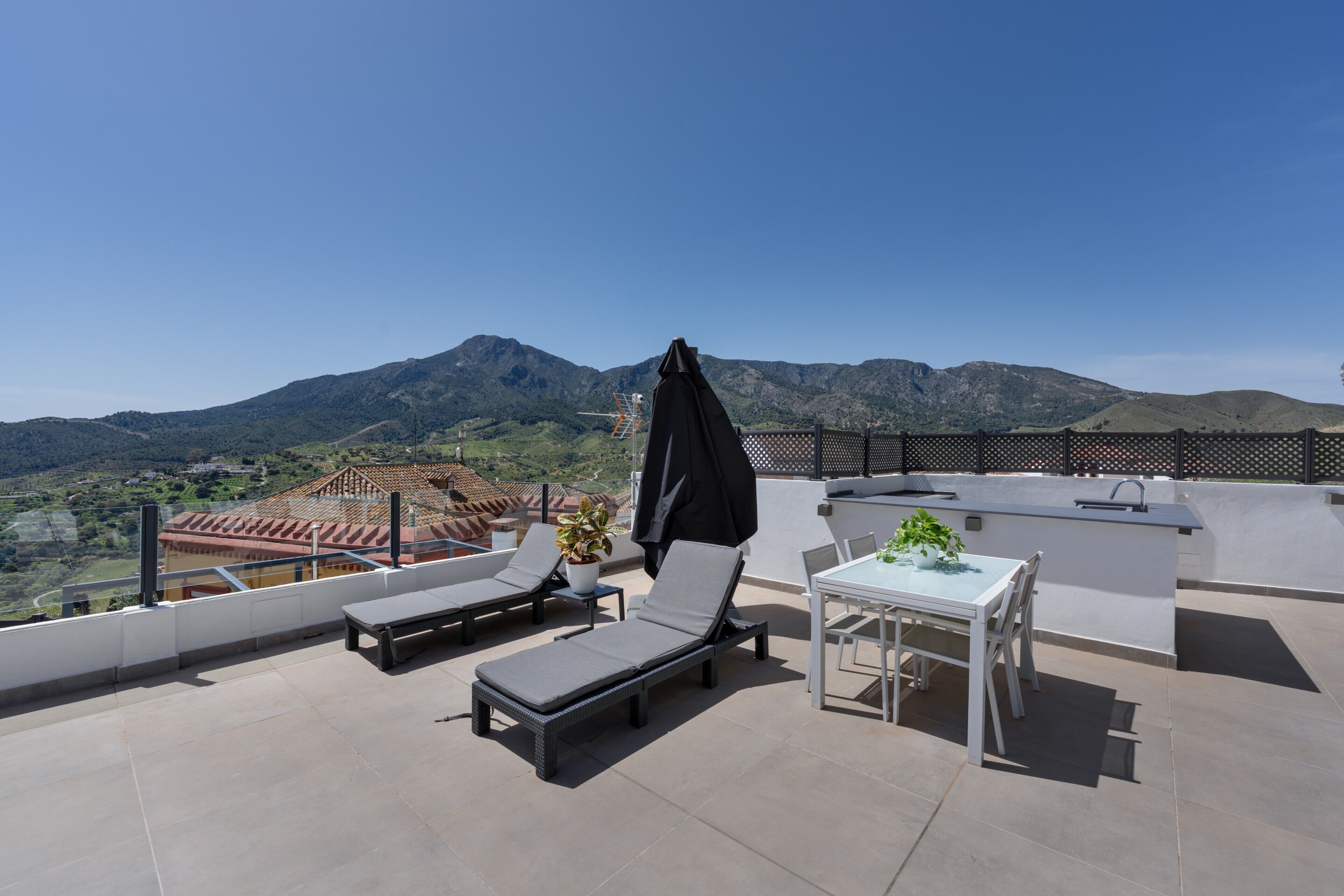 Terraço no topo do prédio com duas espreguiçadeiras, uma mesa com cadeiras, um vaso pequeno com planta e um guarda-sol preto.