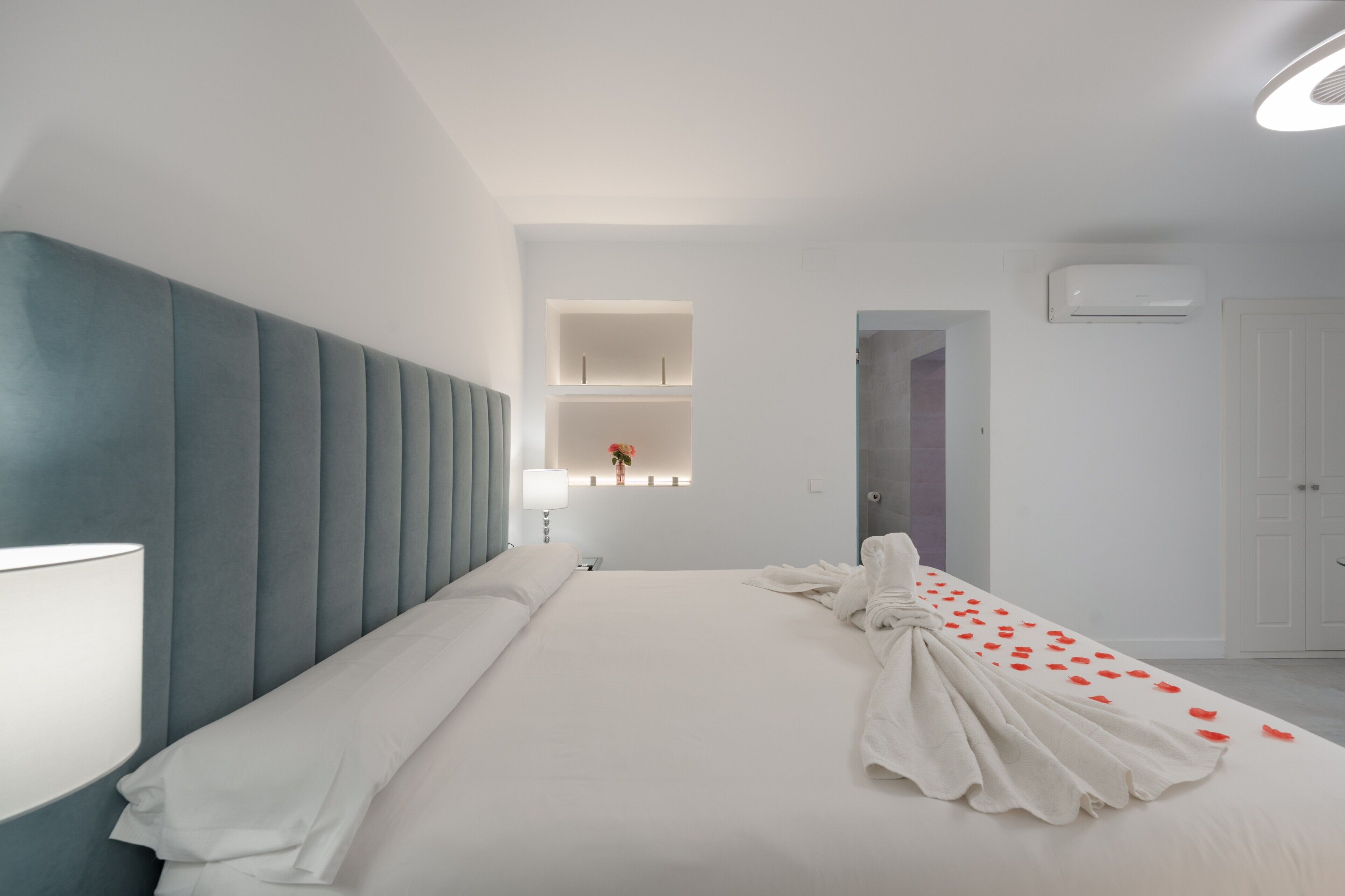 A modern hotel room with a large bed, a headboard, a bedside table, a lamp, a wall-mounted shelf with a vase, and an air conditioning unit.