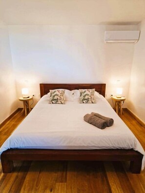 A single bed with a wooden frame, white bedding, two pillows with a leaf pattern, and a rolled towel on the bed. There are two bedside tables with lamps, and an air conditioning unit mounted on the wall above the bed. - La Case Bleue (Les Avirons)