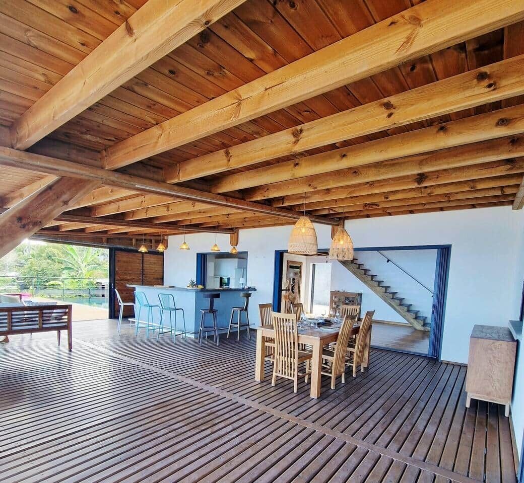 An open-plan living area with a wooden ceiling, a dining table with chairs, a kitchen area, and a staircase leading to an upper level.
