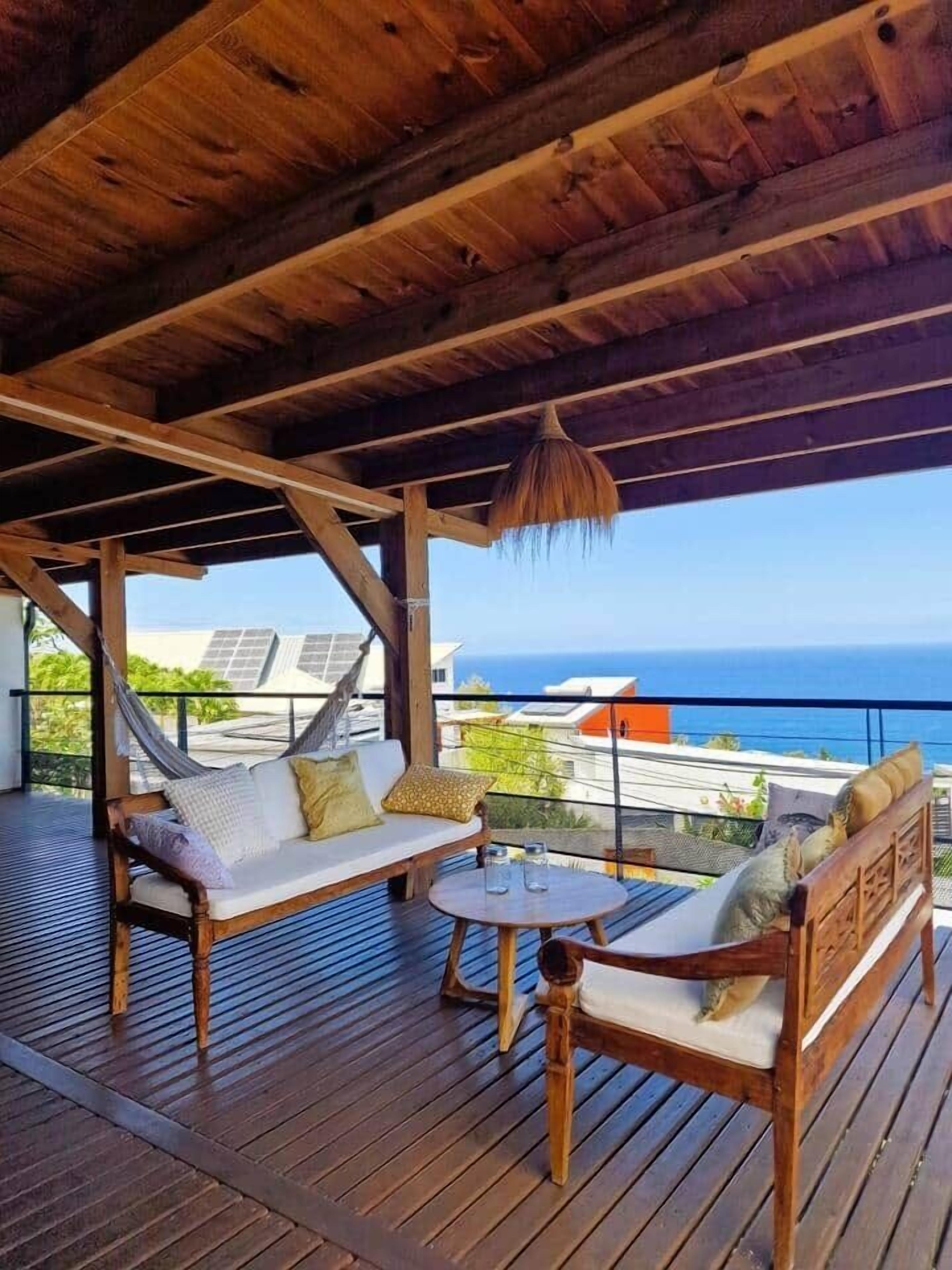 Une terrasse en bois avec un hamac, une table ronde et un canapé avec des coussins.