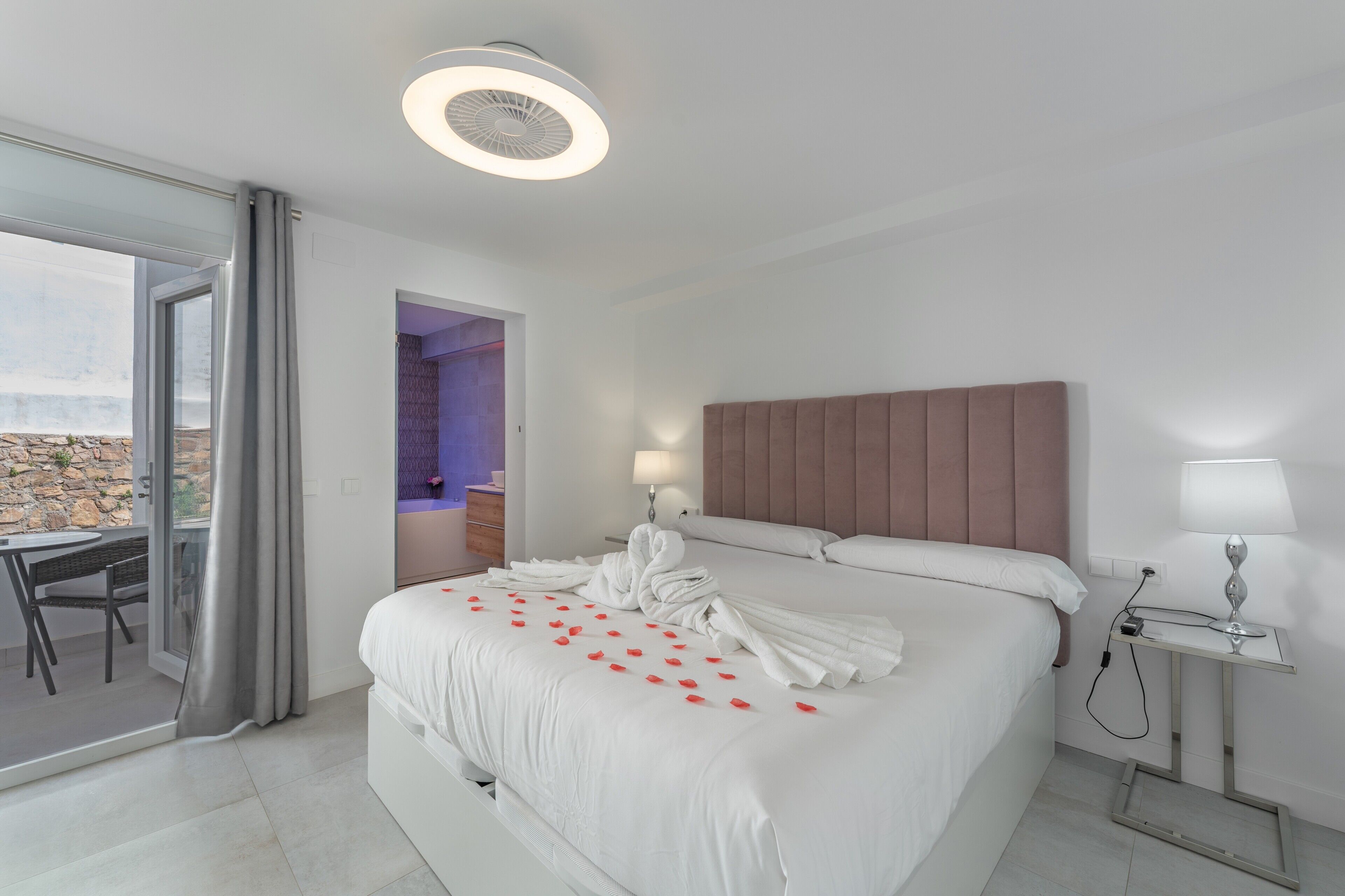 A hotel room with a bed, a bedside table with a lamp, a ceiling light, and a view through a glass door to an outdoor area.
