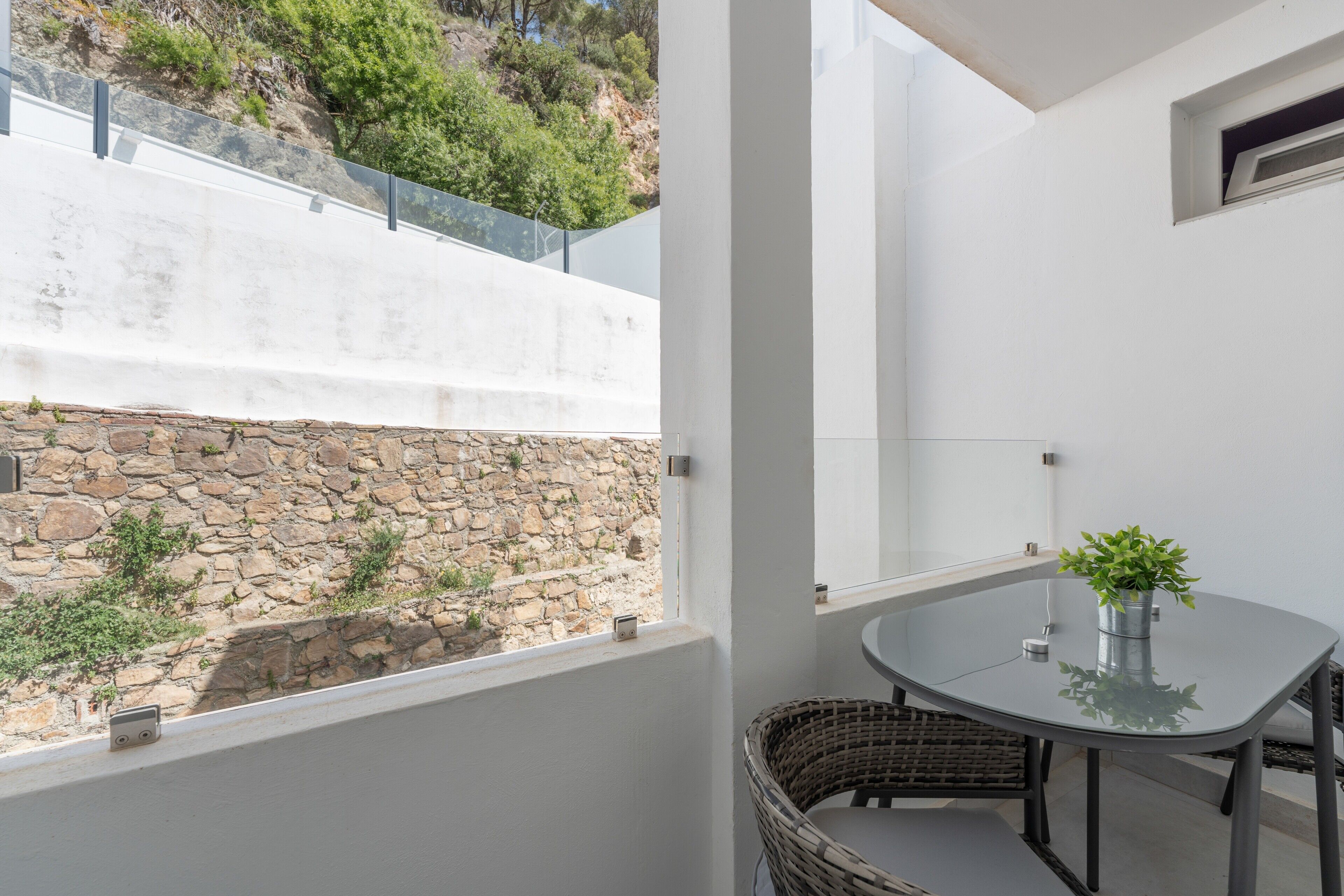 Un balcón con pared de piedra, una silla de mimbre, una mesa de vidrio y una planta en maceta.