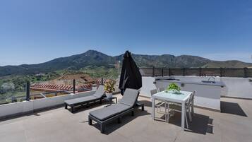 A rooftop terrace with two sun loungers, a table with chairs, a small potted plant, and a black umbrella.