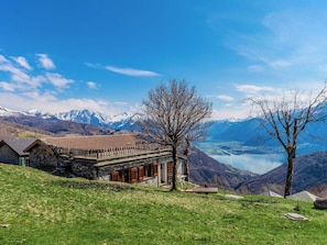 Ein Gebäude aus Stein mit einem geneigten Dach, einer Holzterrasse und Blick auf einen See und Berge.