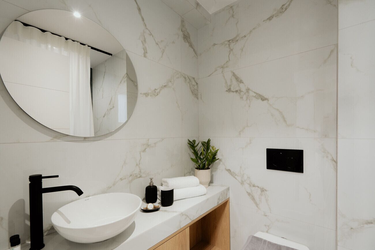 A bathroom with a round mirror, a white sink, a black faucet, a potted plant, and a towel rack.