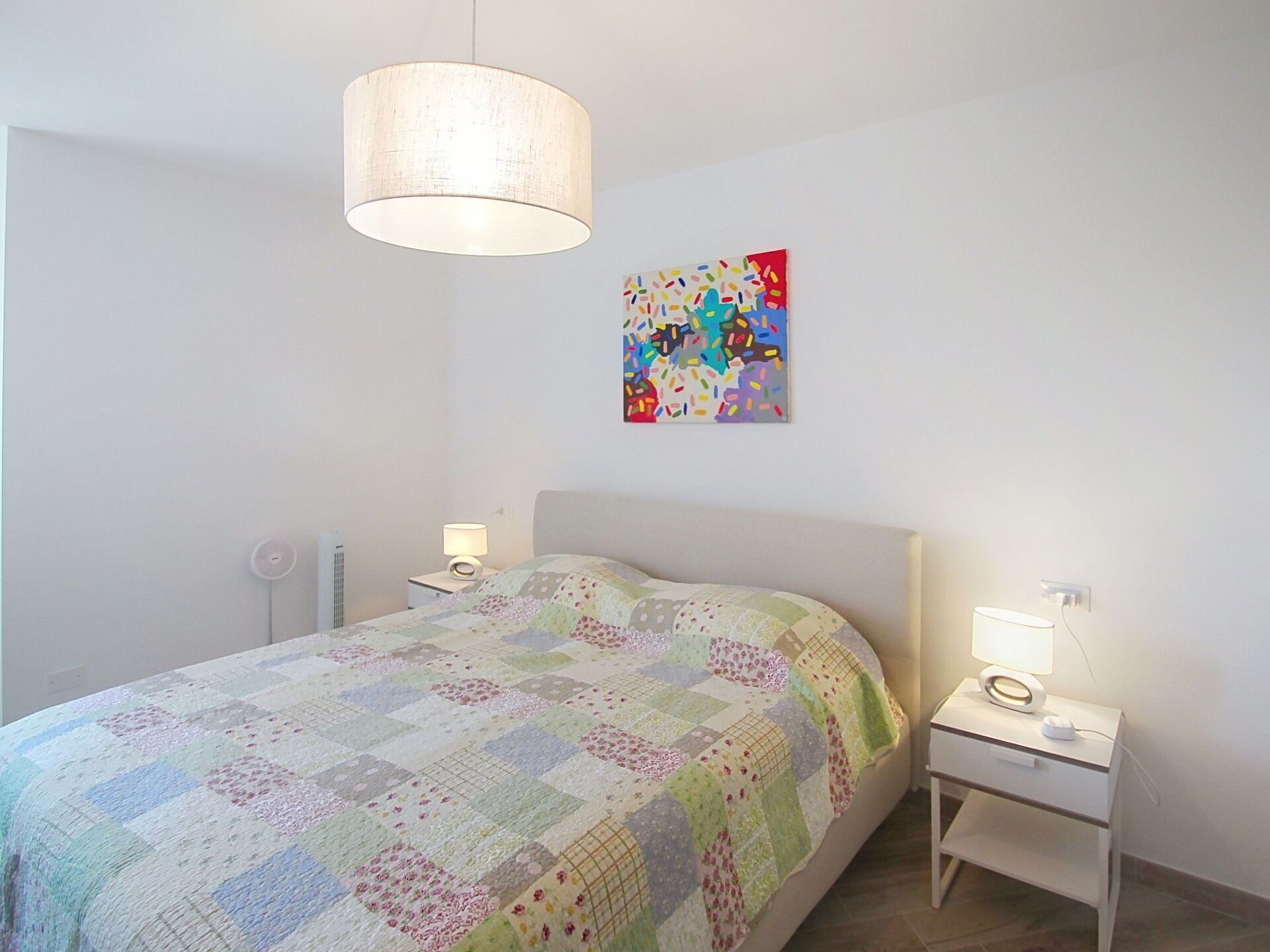 A bedroom with a bed, two bedside tables, a hanging lamp, a wall-mounted picture, and a radiator.