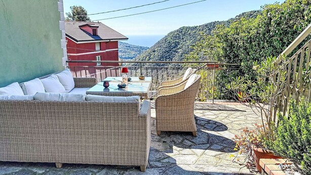Eine Terrasse mit Korbmöbeln, einem Tisch mit roter Lampe und Blick auf ein rotes Gebäude und Hügel.