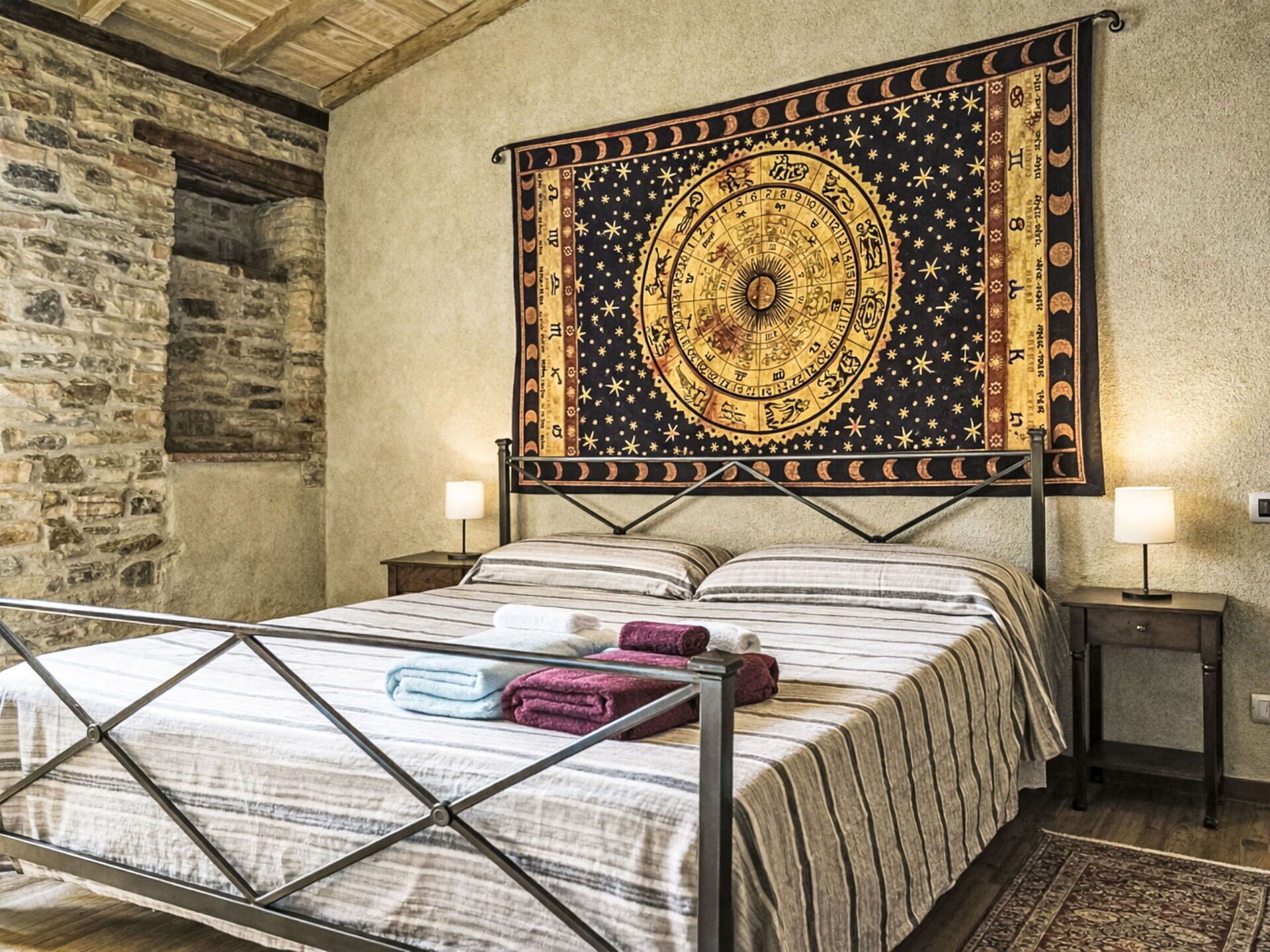 Una cama con estructura metálica, un tapiz grande con símbolos zodiacales y dos lámparas de noche.