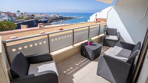 A balcony with wicker furniture, a potted plant, and a view of the sea and buildings.