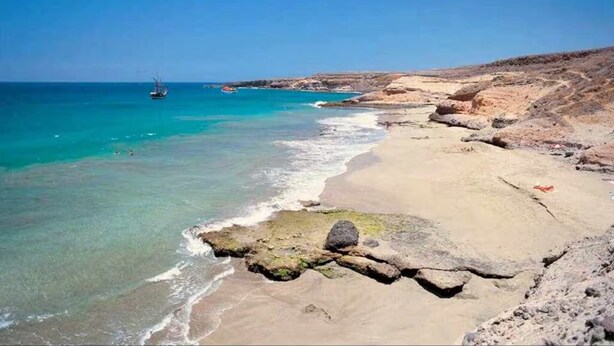 A coastal beach with clear water, rocky terrain, and a boat in the distance.