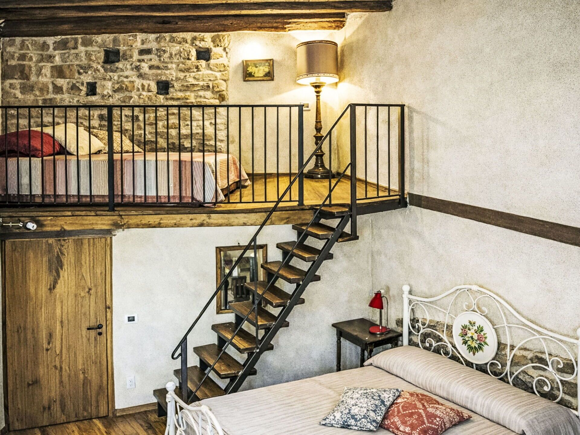 A bedroom with a loft bed, a wooden staircase, a stone wall, and a small table with a lamp.