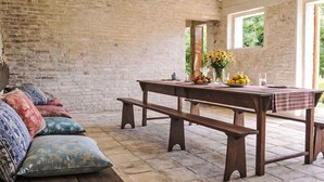 Ein rustikaler Raum mit Backsteinwand, einer Holzbänkl, einem Tisch mit kariertem Tuch und einer Couch mit gemusterten Kissen.