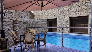 An outdoor pool area with a brick wall, a table with chairs, and a large umbrella. - SIX DIAMOND HOTEL OPEBI (Lagos)