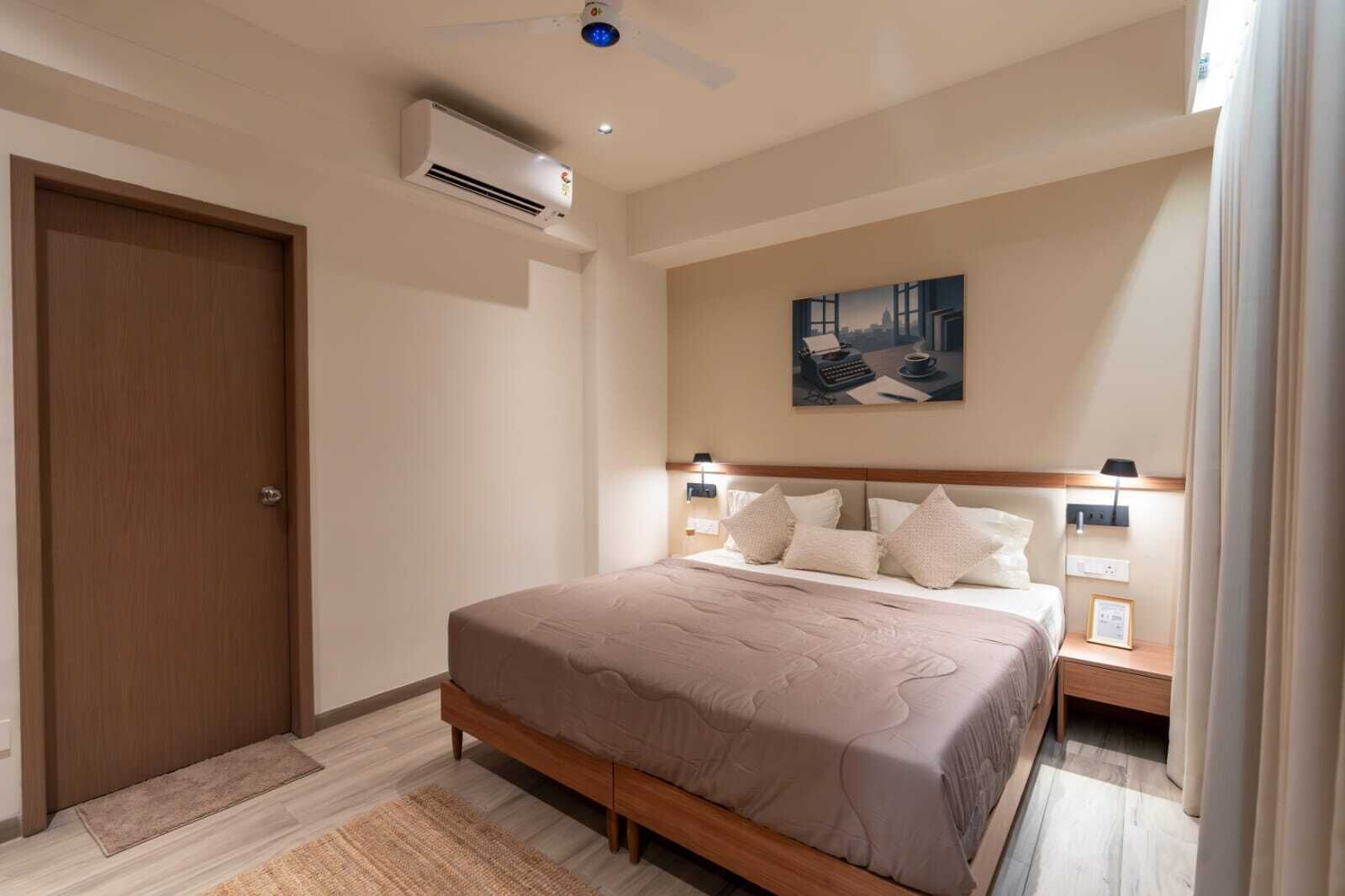 A bedroom with a bed, a nightstand, a wall-mounted air conditioner, a door, and a painting on the wall.