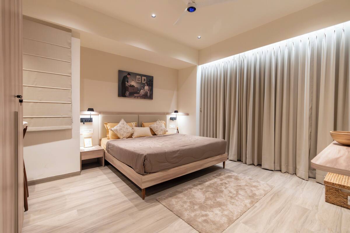 A bedroom with a bed, bedside tables, a nightstand, a painting, a ceiling fan, and a window with curtains.