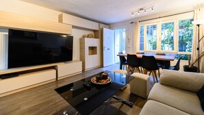 A modern living room with a flat-screen TV, a glass coffee table, a sofa, a dining area with chairs, and a large window with a view of greenery. - (SAB52) Spacious 3-bedroom villa with terrace (Madrid)