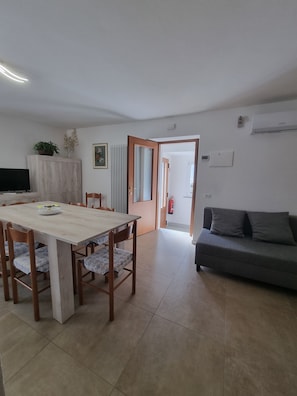 A living room with a sofa, a dining table with chairs, a TV, and a kitchen area with cabinets and a plant. - Well House (Vercana)