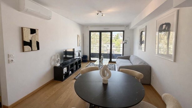 Un salon moderne avec une table à manger noire, un canapé gris et un téléviseur posé sur un meuble noir. Une grande fenêtre offre une vue sur l’extérieur.