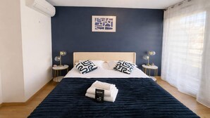 Un lit bien fait, recouvert d’une couverture bleu foncé, deux lampes de chevet, une œuvre d’art encadrée au mur et une fenêtre avec des rideaux légers.