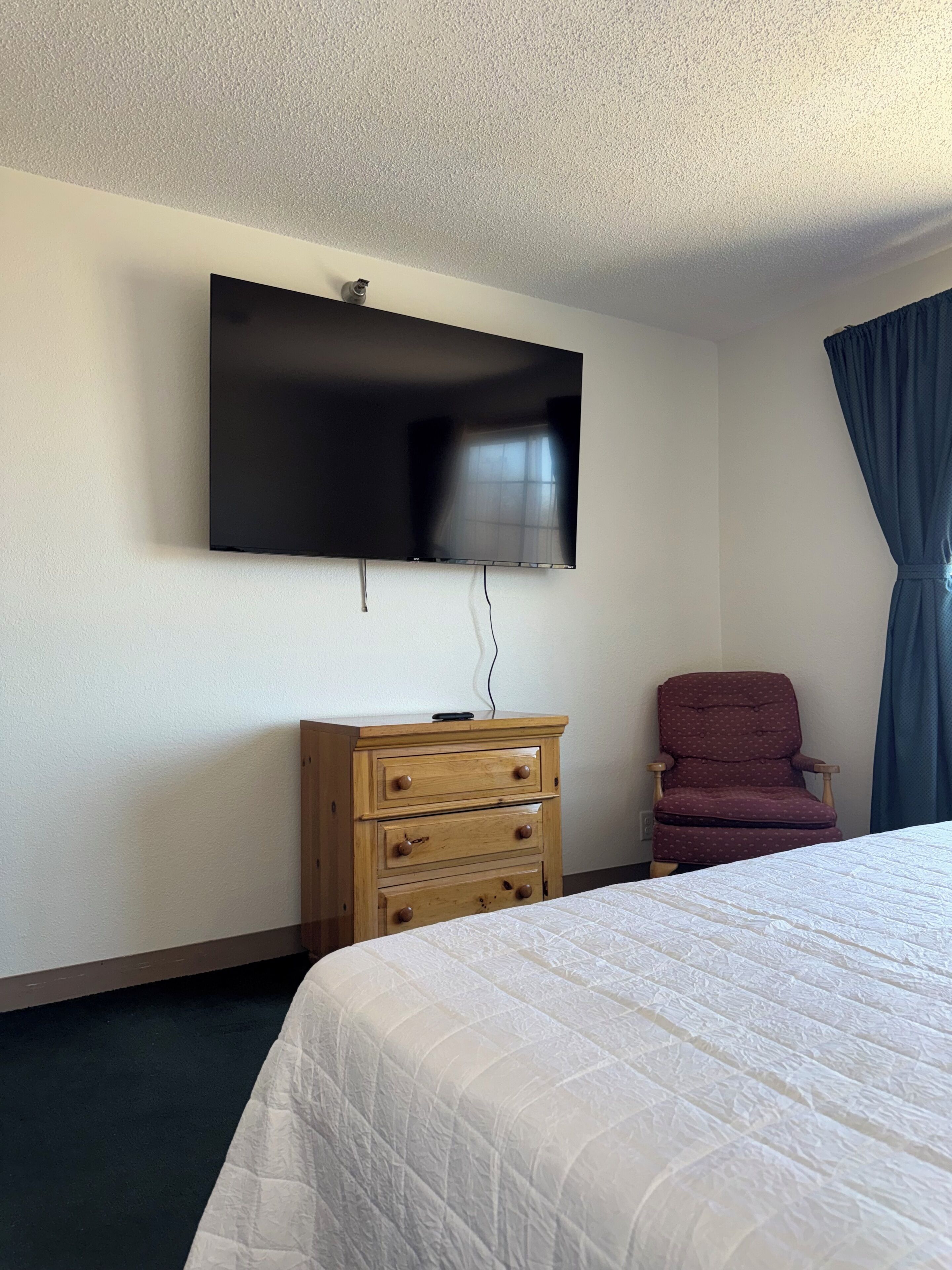 A hotel room with a bed, a wooden dresser, a television mounted on the wall, and a chair.