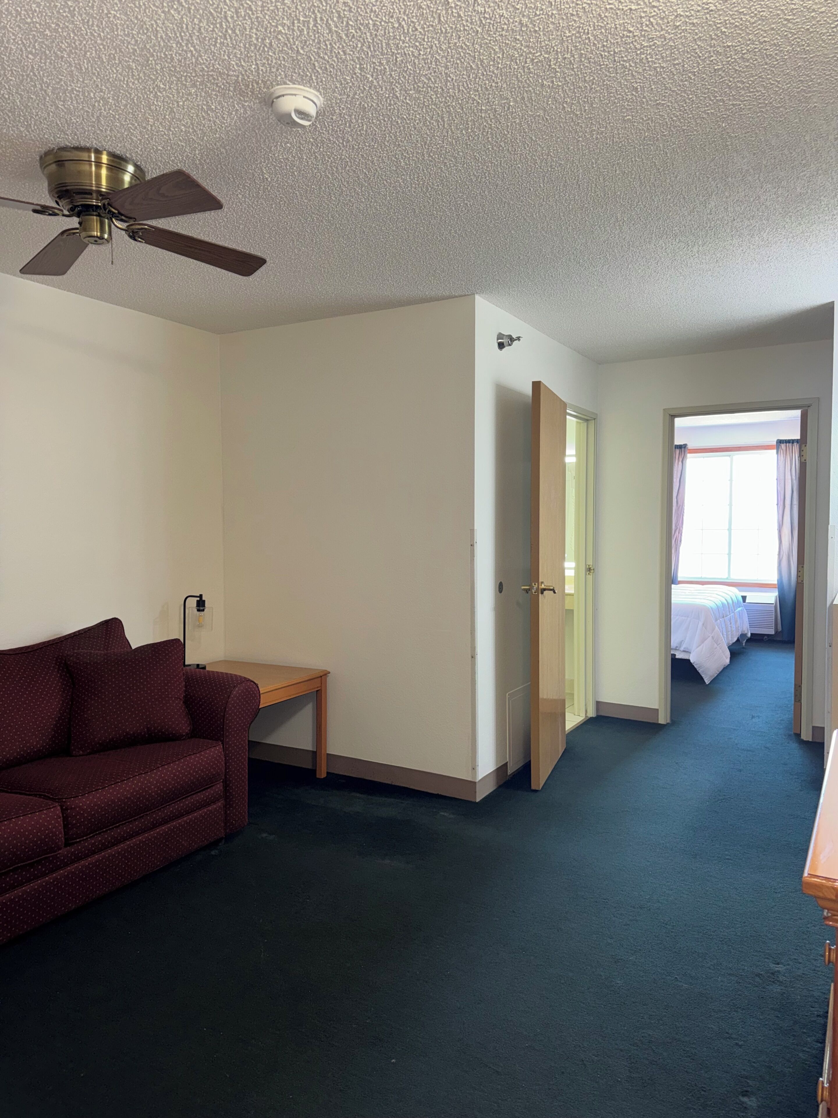 A hotel room with a ceiling fan, a red sofa, a small table, a door leading to a bedroom, and a window with curtains.