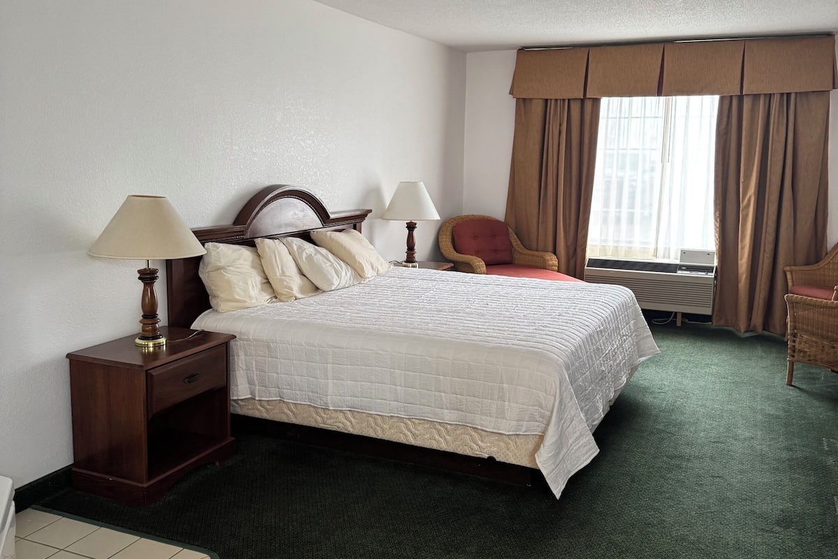A hotel room with a bed, two bedside tables, a chair, a window with curtains, and an air conditioning unit.