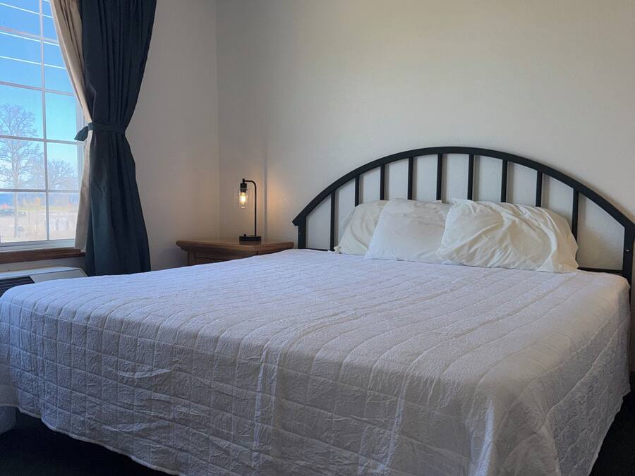 A neatly made bed with a white quilt, a bedside table with a lamp, and a window with curtains.