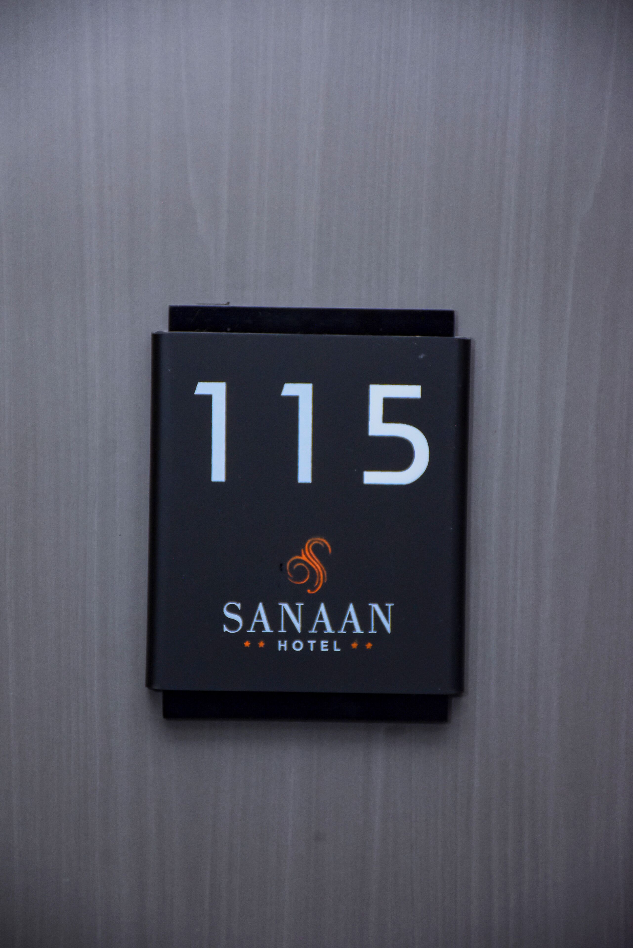 Un letrero negro con el número 115 y el nombre 'Sanaam Hotel' sobre una pared gris.