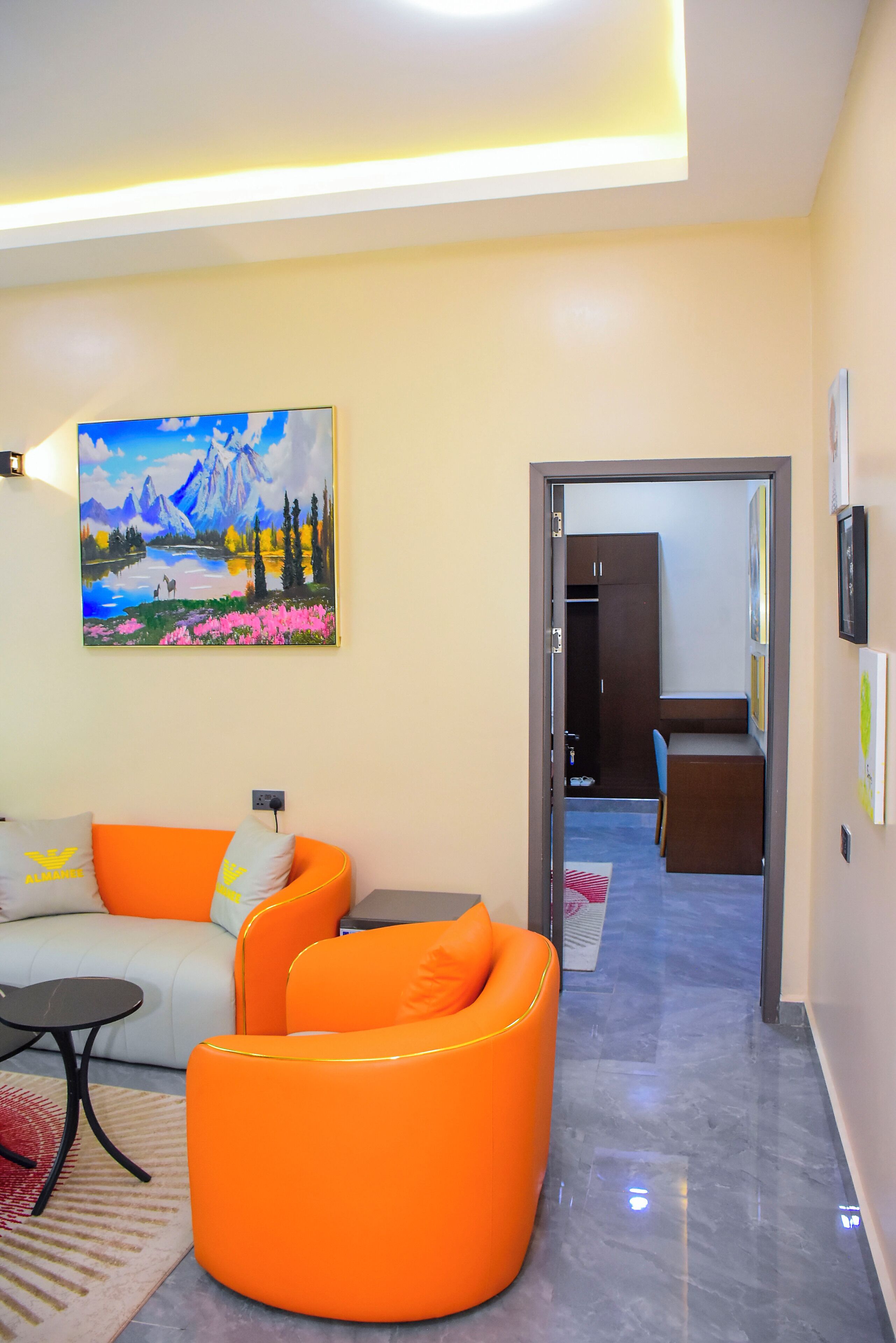 Uma sala de estar moderna com um sofá, poltronas laranja, um quadro e um corredor que dá para uma cozinha.