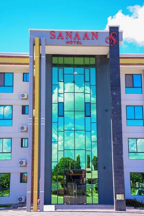 A modern hotel with a large glass facade, a prominent sign reading 'SANAAN HOTEL,' and a reflection of the sky and trees.