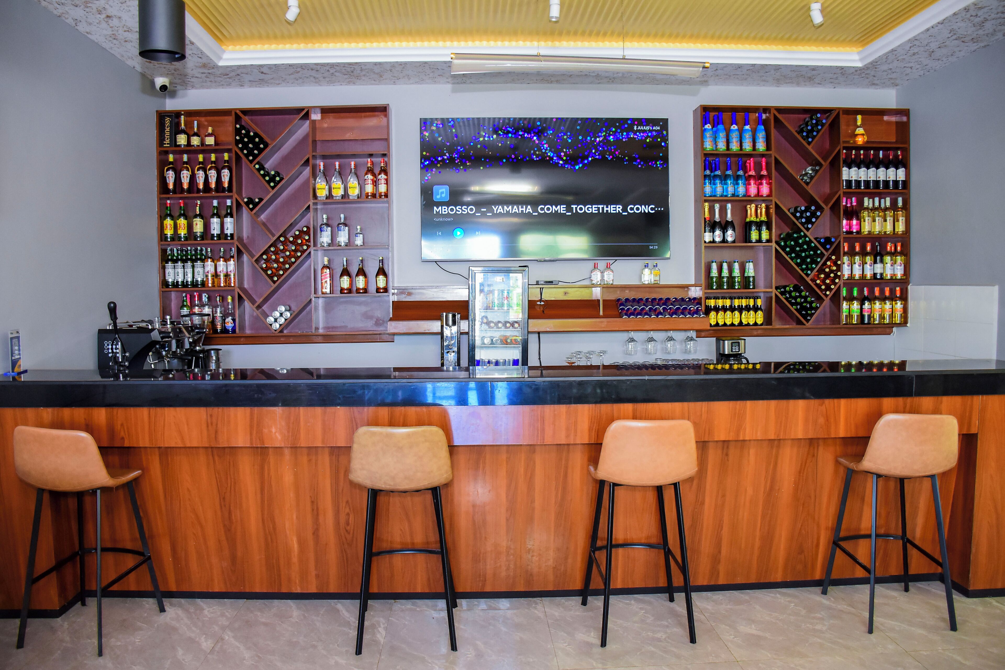 Una zona de bar con una pantalla de televisión, estantes de madera con botellas y tres taburetes de bar.