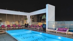 Un espace piscine aménagé sur le toit, avec des chaises longues et un canapé, offrant une vue panoramique sur la ville la nuit.