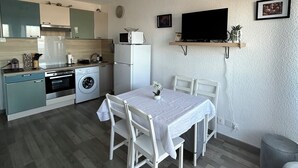 Una cocina compacta con microondas, horno y lavadora; un comedor con mesa y sillas; un televisor de pantalla plana montado en la pared; y una pequeña zona de estar con un banco acolchado.
