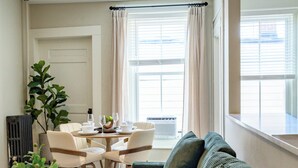 Una sala de estar con un sofá verde, una mesa de comedor con sillas, una planta en maceta y un radiador.