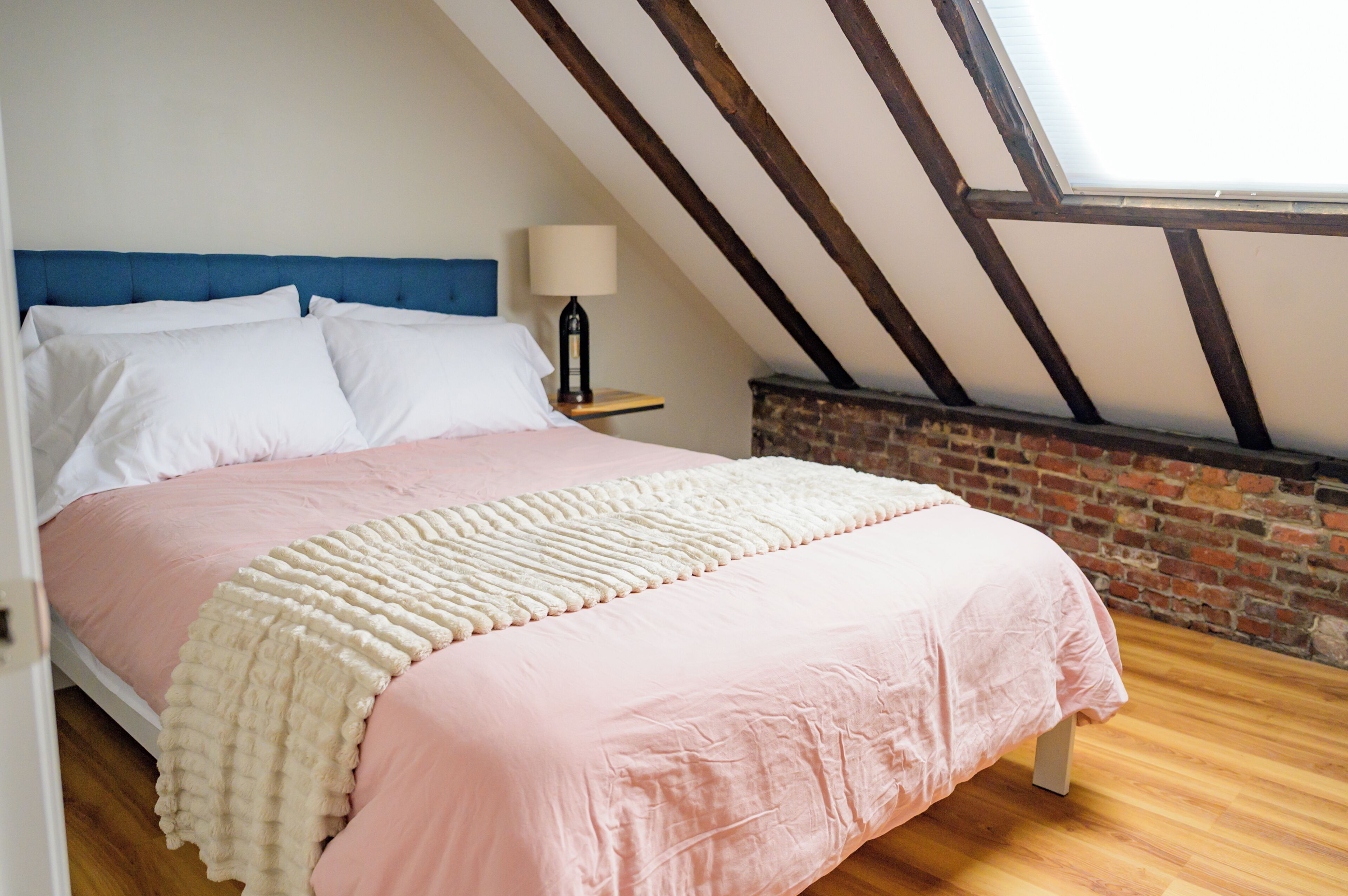 A bedroom with a sloped ceiling, a brick wall, a bed with pink bedding, a white throw blanket, and a nightstand with a lamp.