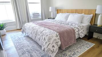 Un dormitorio con cama, mesitas de noche, una planta en maceta y una ventana con cortinas.
