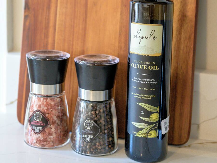 Una botella de aceite de oliva extra virgen, dos recipientes de granos de pimienta y una tabla de cortar de madera.