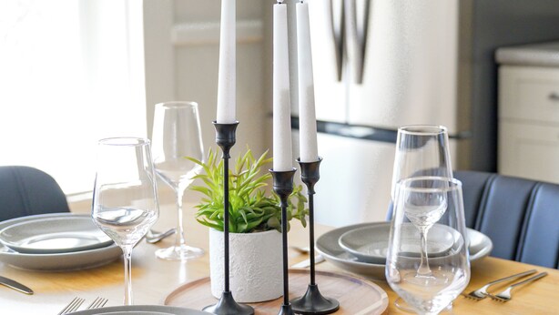 Una mesa de comedor dispuesta con platos, vasos, cubiertos y velas, con un refrigerador y armarios al fondo.
