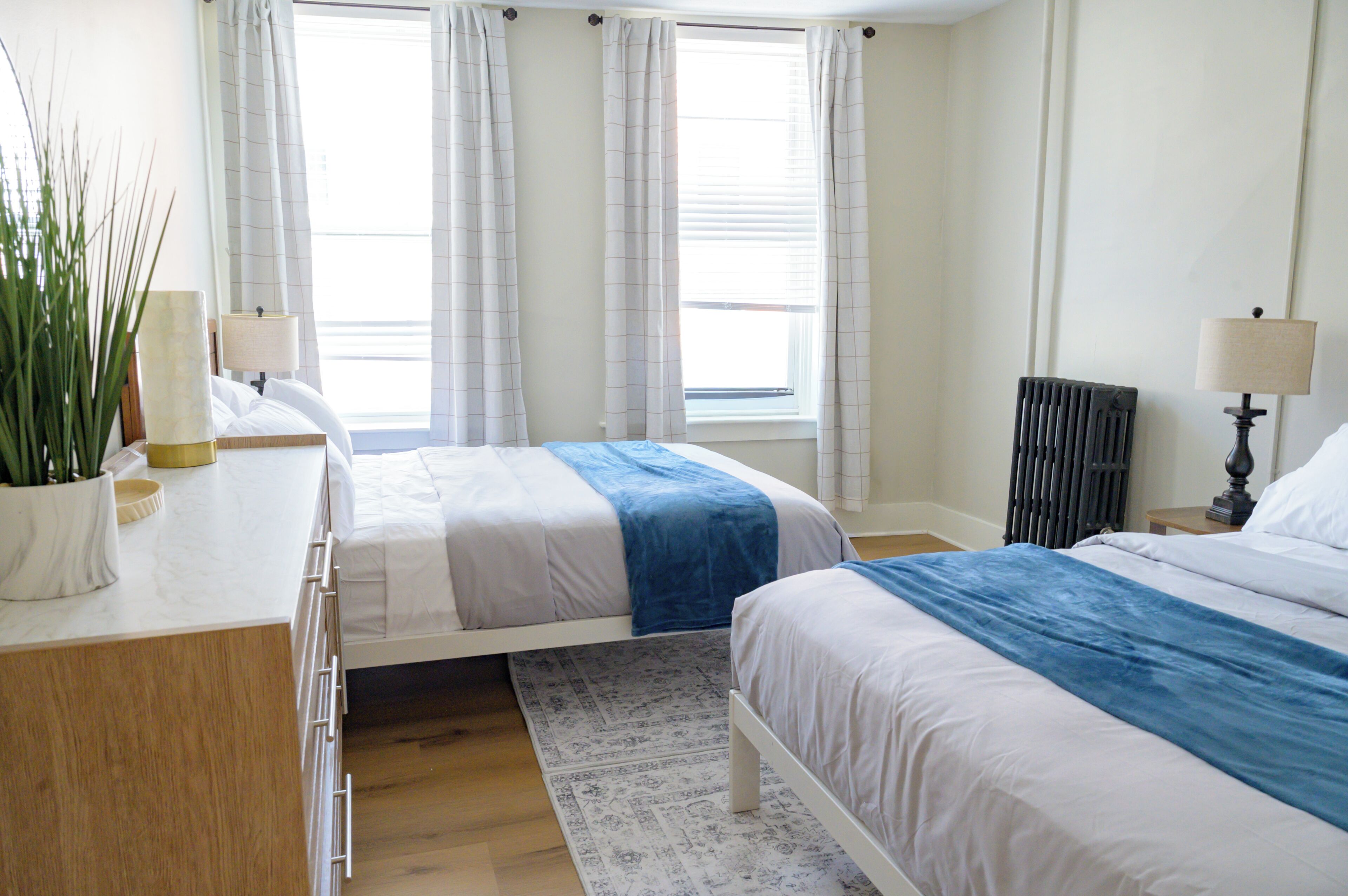 A bedroom with two beds, a radiator, a bedside table with a lamp, a potted plant, and a window with curtains.