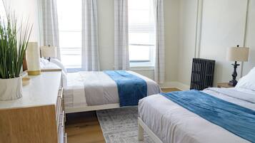 A bedroom with two beds, a radiator, a bedside table with a lamp, a potted plant, and a window with curtains.